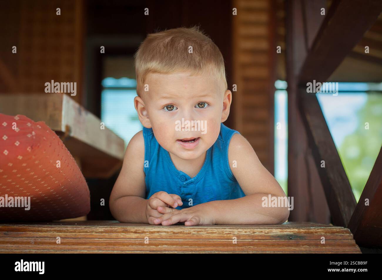 Un bambino con i capelli corti si appoggia curiosamente in avanti su una superficie di legno. La luce solare filtra nell'interno, concentrandosi sull'espr. Del bambino Foto Stock