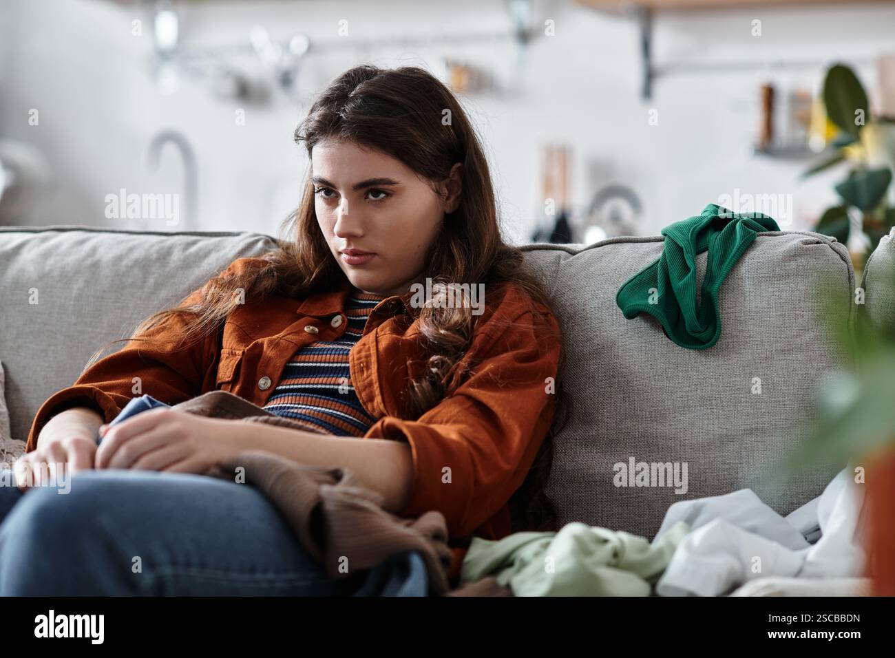 Una donna siede tranquillamente su un divano, circondata da vestiti disordinati, mostrando la sua tristezza interiore. Foto Stock
