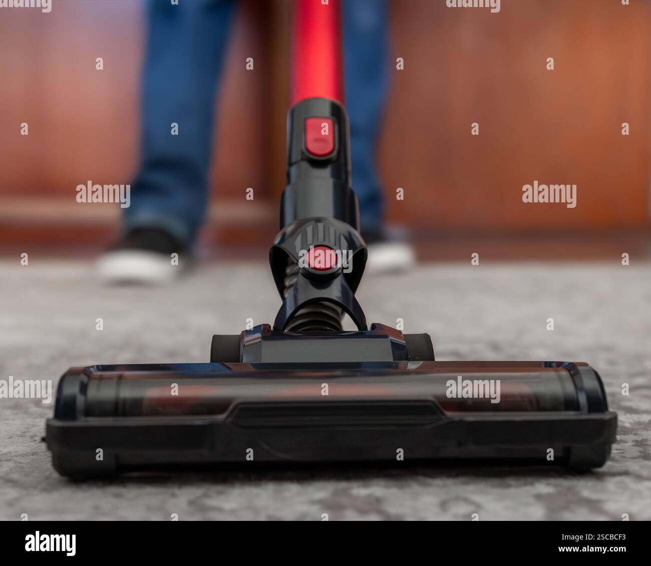 Primo piano ad angolo basso di un aspirapolvere cordless utilizzato su un pavimento con moquette. La composizione dinamica e lo sfondo sfocato conferiscono un'atmosfera piacevole Foto Stock