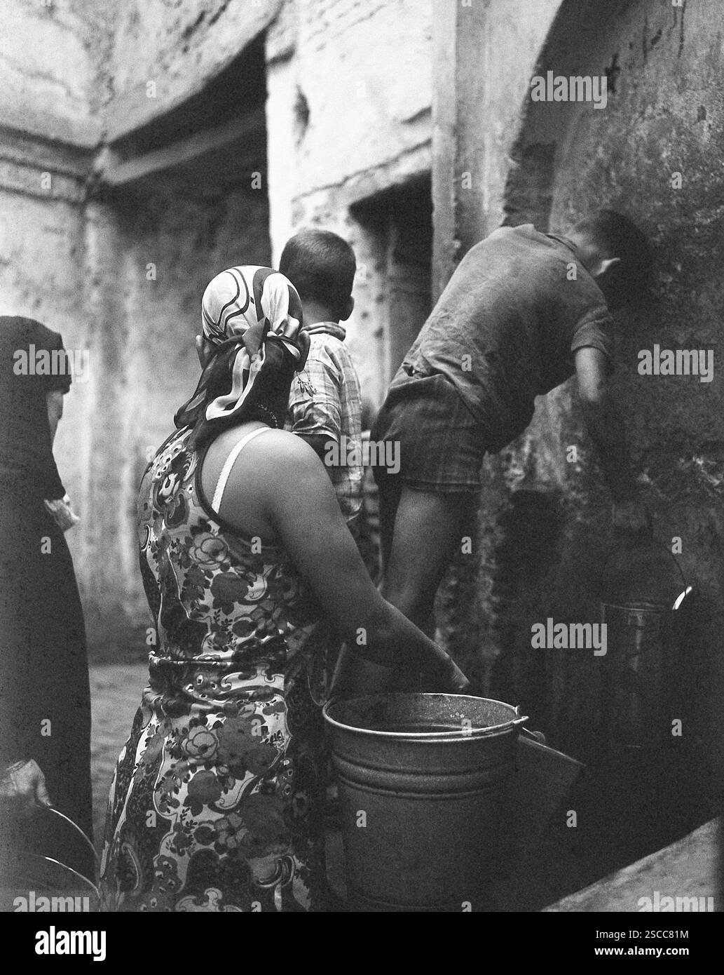 I bambini e le donne con headscarfs e benne in corrispondenza di un foro per l'acqua in Meknes in Marocco. Foto Stock