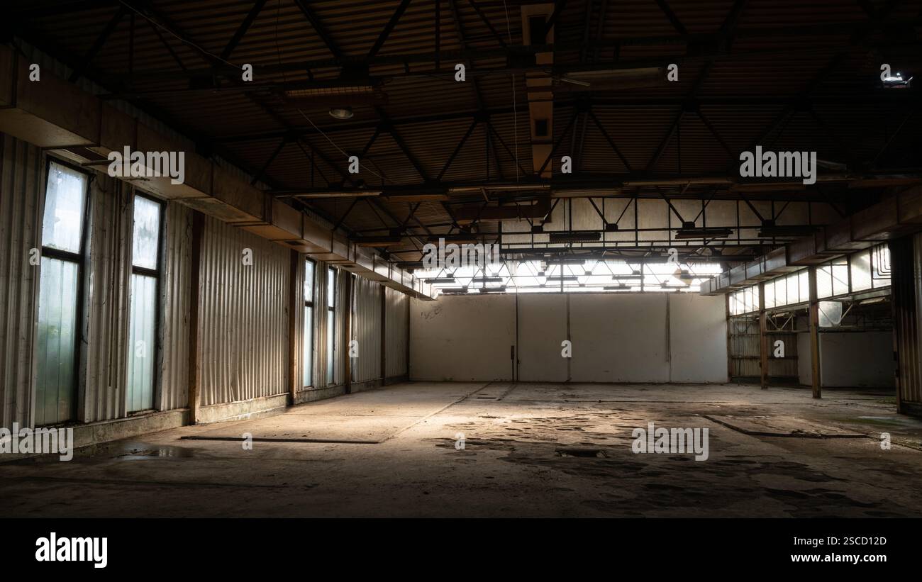 Interno di una sala industriale vuota abbandonata, realizzata in lamiere e pannelli di vetro illuminati dal sole attraverso la finestra Foto Stock