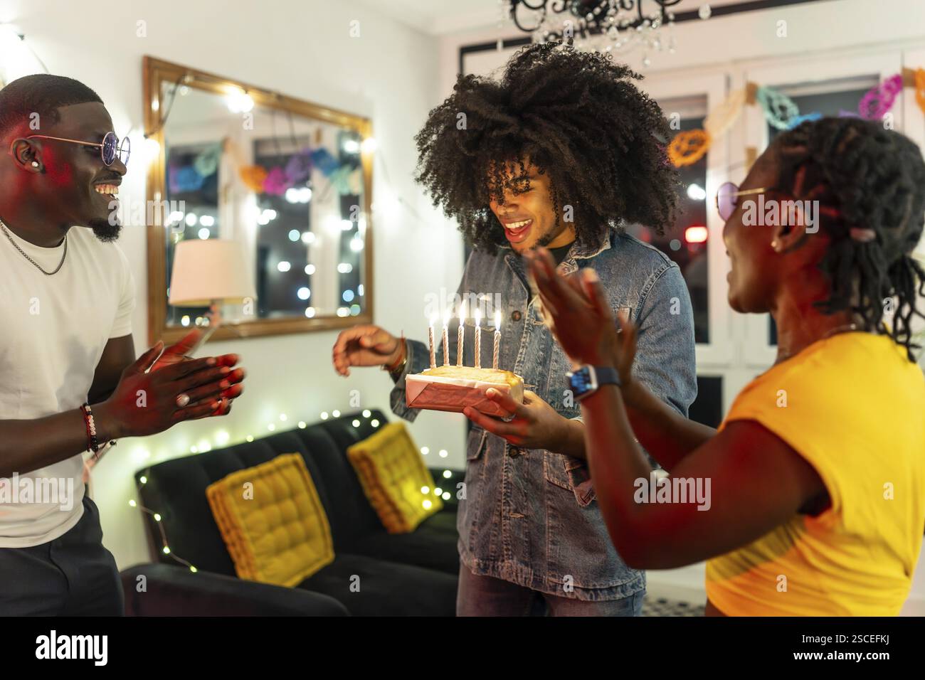 Amici multietnici che cantano una canzone di compleanno a un amico che tiene una torta festeggiata a casa Foto Stock