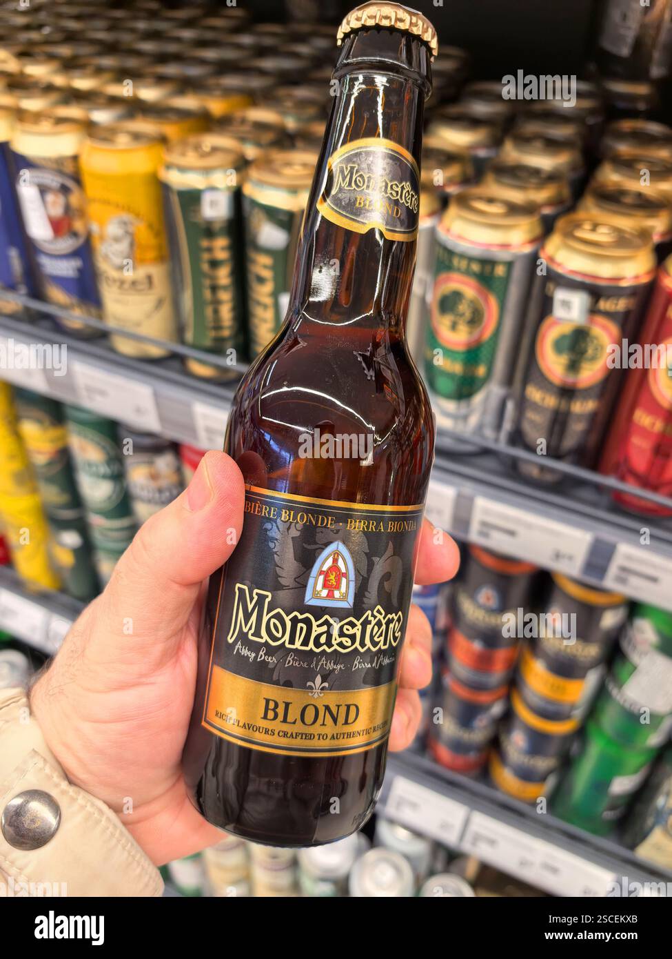 Bobruisk, Bielorussia - 2 febbraio 2025: Una mano tiene una bottiglia di birra artigianale bionda in un corridoio ben rifornito. Varie altre birre in scatola e in bottiglia a. Foto Stock