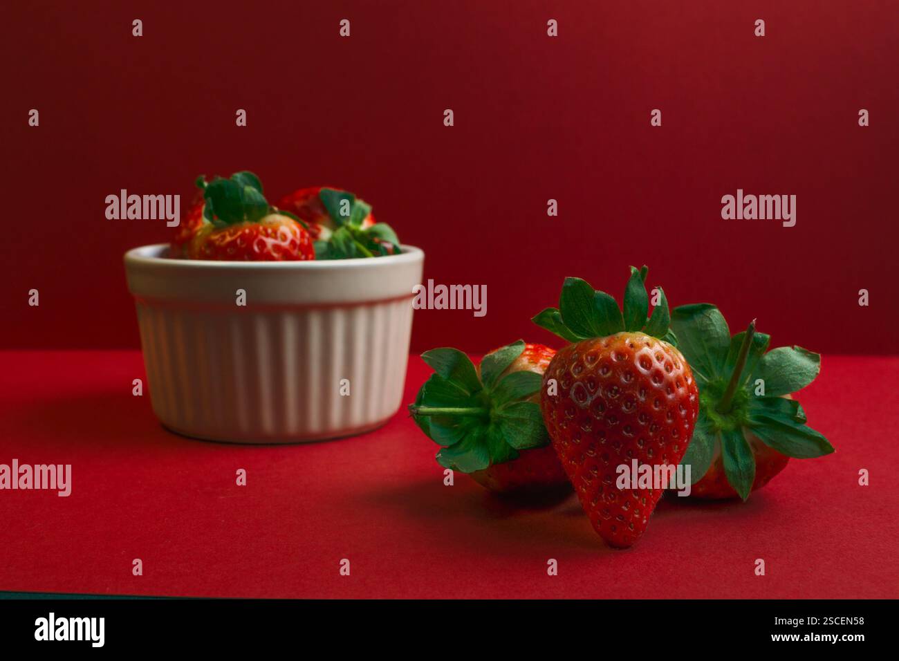 Una ciotola di fragole è su sfondo rosso. Ci sono tre fragole sul tavolo. La ciotola è bianca Foto Stock