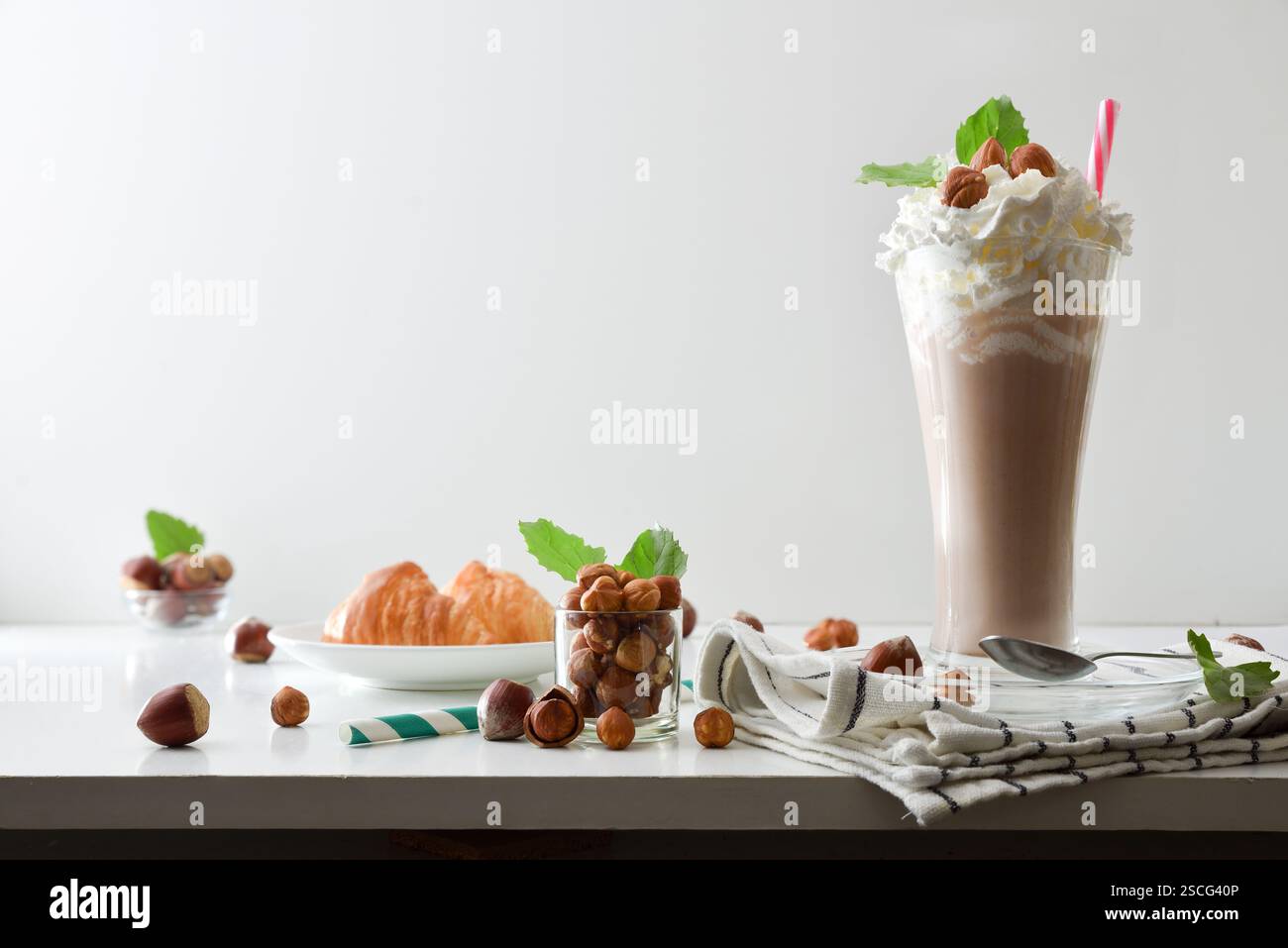 Spuntino sul tavolo bianco con bicchiere di frullato di nocciole con panna e croissant decorati con ciotole con nocciole e foglie e ba bianca isolata Foto Stock