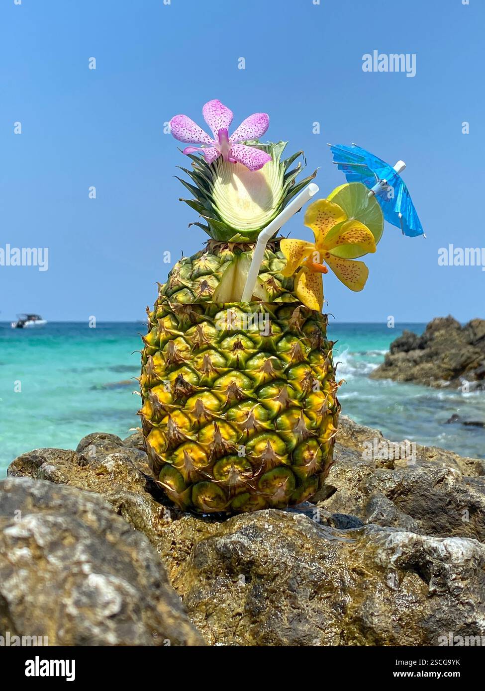 Un cocktail fresco a base di ananas decorato con orchidee rosa e gialle e lime con ombrellone in piedi sulla spiaggia, mare blu sullo sfondo Foto Stock