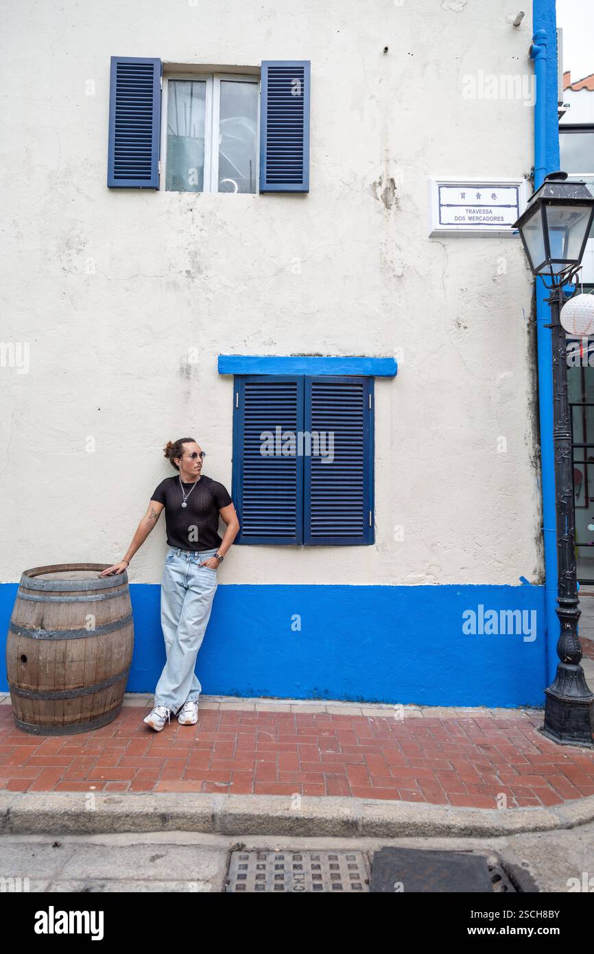 Un uomo, appoggiato al muro, accanto a botte di legno e finestra blu Foto Stock