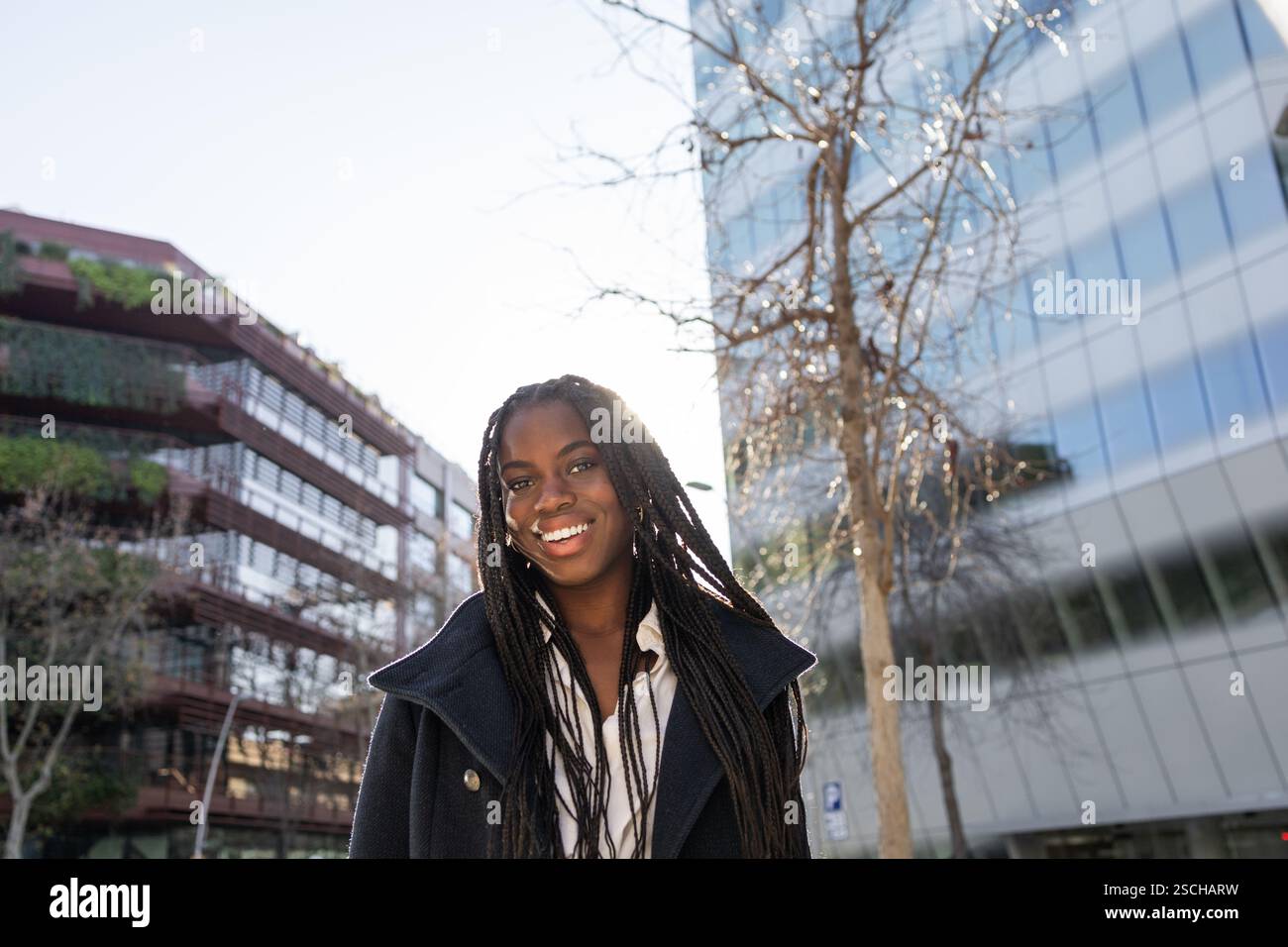 Dal basso, donna d'affari afroamericana sorridente con trecce che si gode una pausa all'esterno dei moderni edifici di uffici, mostrando positività e fiducia. Foto Stock