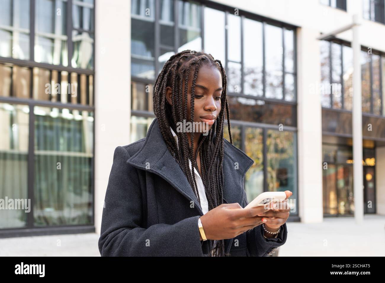 Una giovane donna afroamericana con trecce, vestita con un cappotto da lavoro, sta usando uno smartphone mentre si trova all'aperto vicino a un edificio moderno. Foto Stock