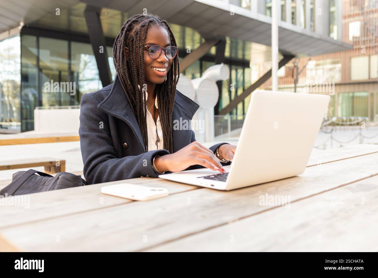 Una giovane donna d'affari afroamericana con trecce lavora su un computer portatile a un tavolo all'aperto in un ambiente informale. Foto Stock