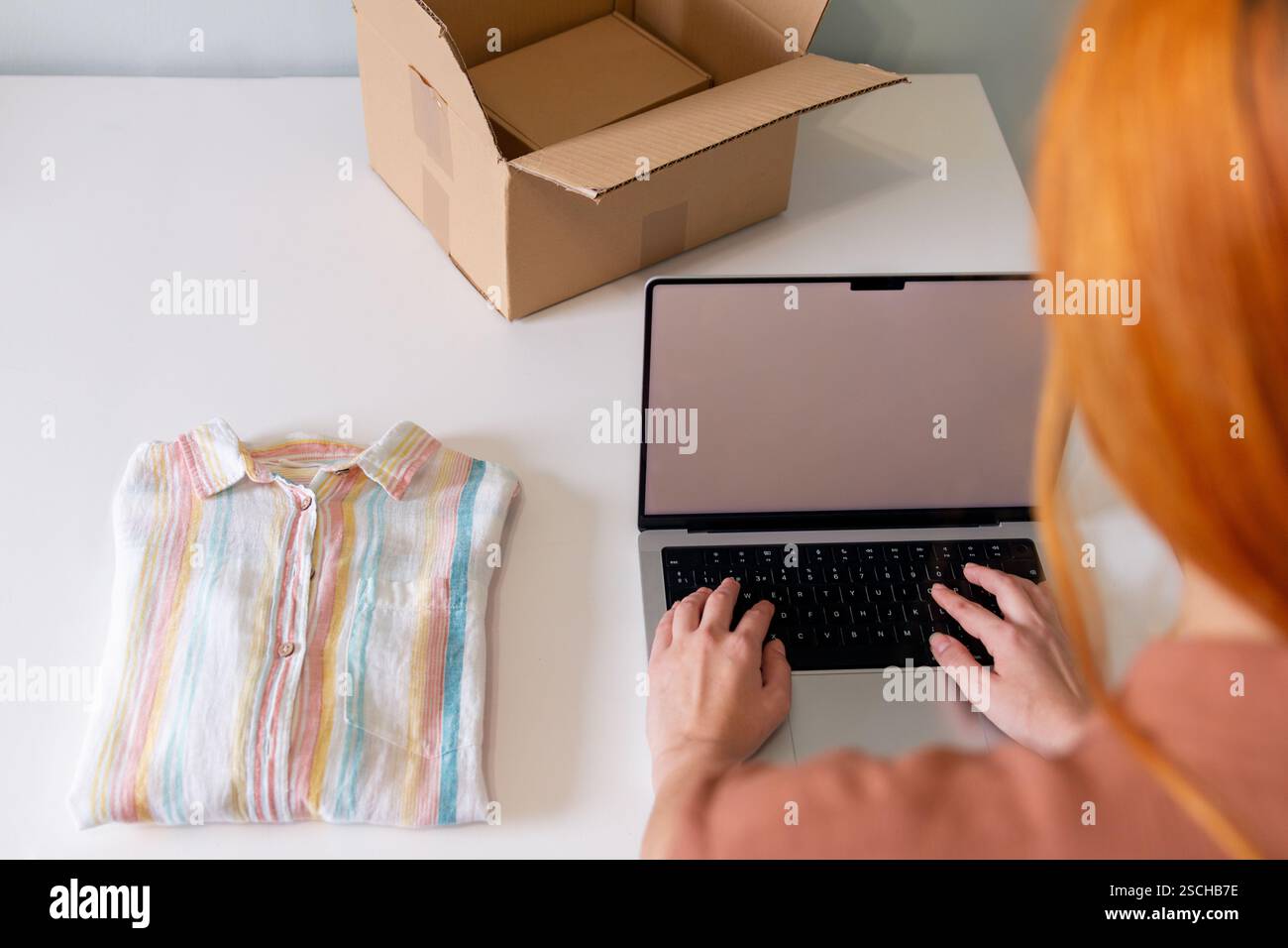 Una giovane donna che organizza abiti in vendita, utilizzando un computer portatile, con una camicia piegata e una scatola aperta nelle vicinanze. La vivace camicia contrasta con la scrivania bianca, e. Foto Stock