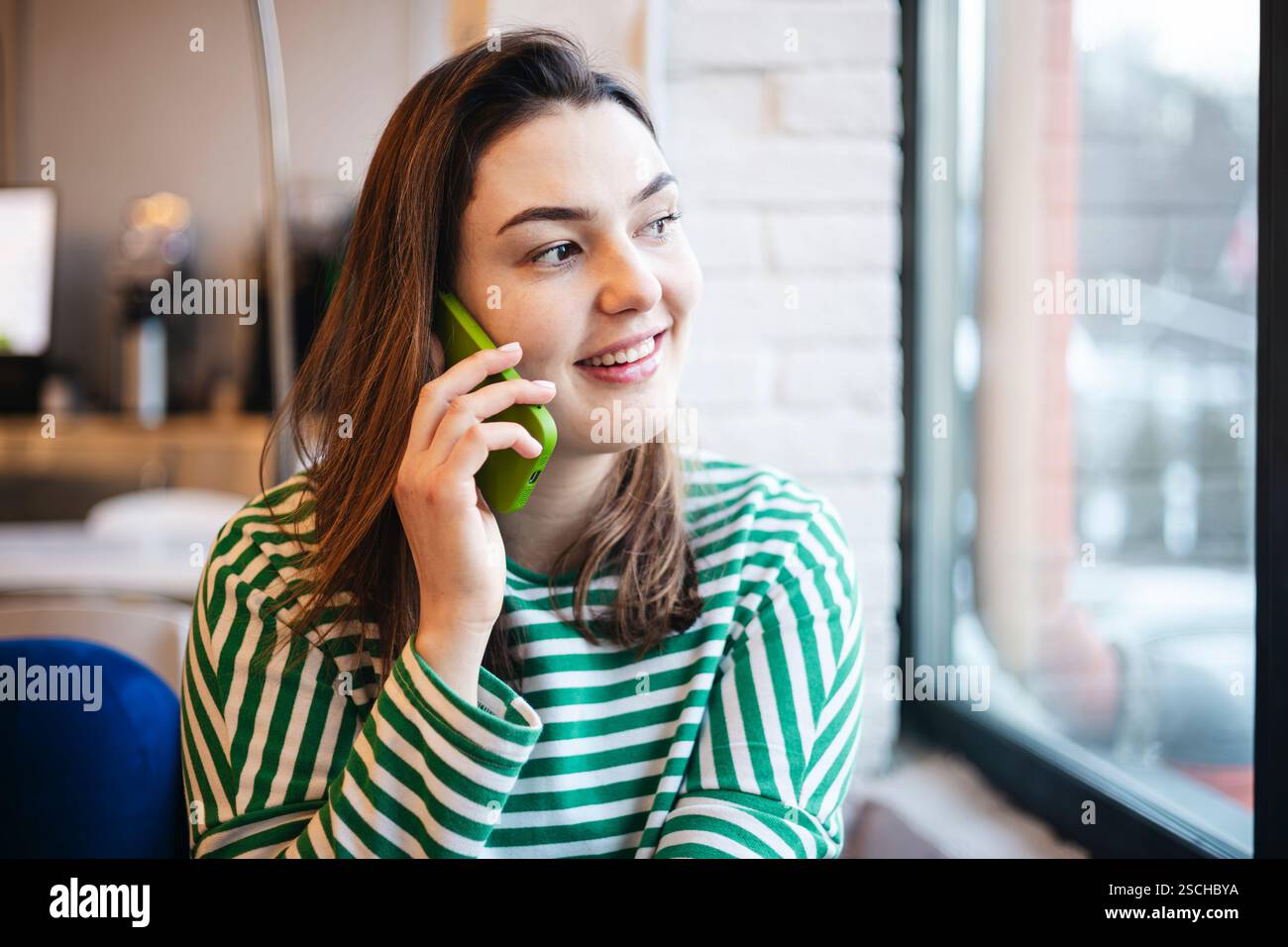 Una donna allegra si gode una pausa caffè in un bar mentre chiacchiera sul suo smartphone, vestito con un top casual a righe. Foto Stock