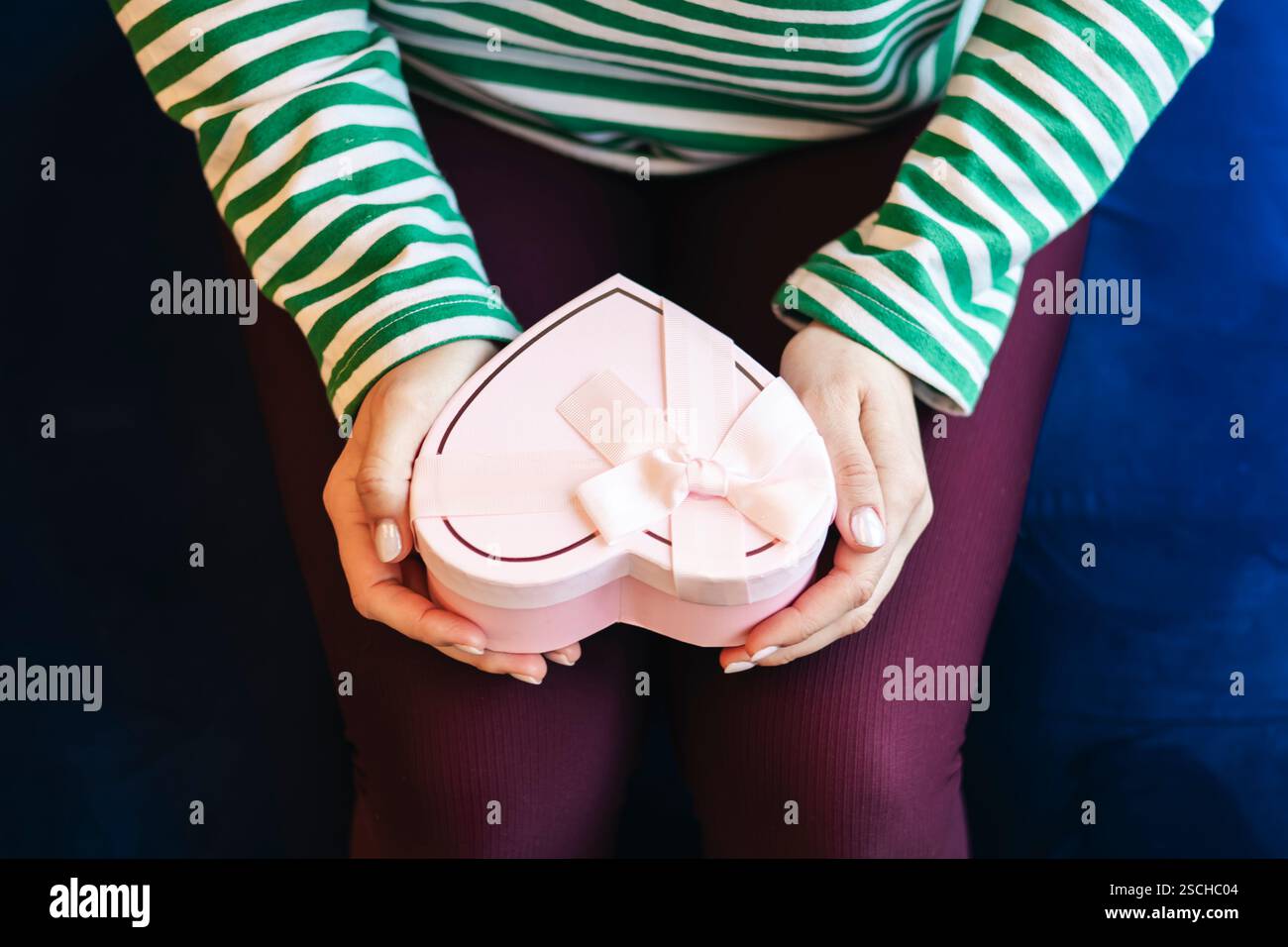 Vista dall'alto, donna irriconoscibile dal taglio corto con una camicia a righe che regge una confezione regalo rosa a forma di cuore. Concentrati sulle mani e sulla scatola, primo piano dettagliato. Foto Stock