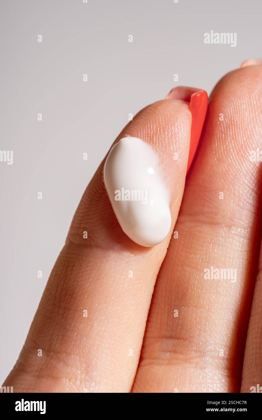 La mano di una donna corta e irriconoscibile con manicure rossa che regge una ricca consistenza di idratante sulla punta delle dita su uno sfondo neutro Foto Stock