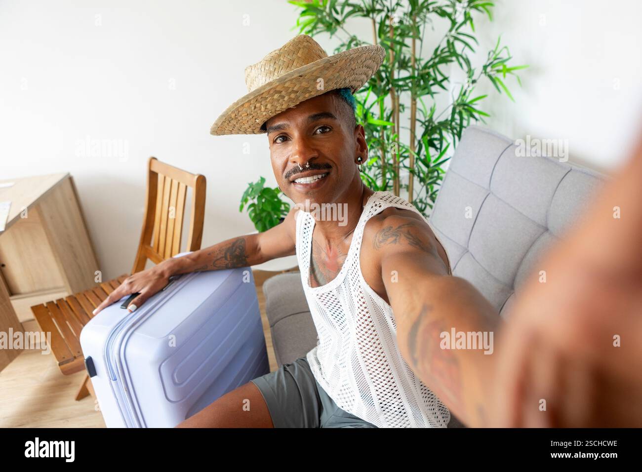 Una persona allegra LGBTQIA+ con una valigia, pronta per le avventure in vacanza. Indossando un cappello di paglia e una maglia a rete, si respirano emozioni e festività Foto Stock