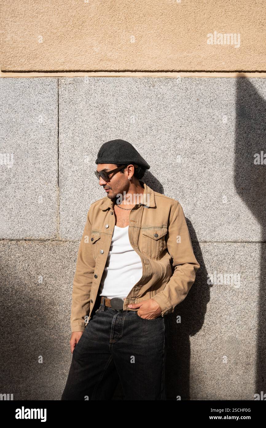 Un uomo si trova di fronte a un muro di pietra, indossando occhiali da sole, un berretto piatto, una giacca beige e una canotta bianca. La sua posa rilassata e il suo abbigliamento donano Foto Stock