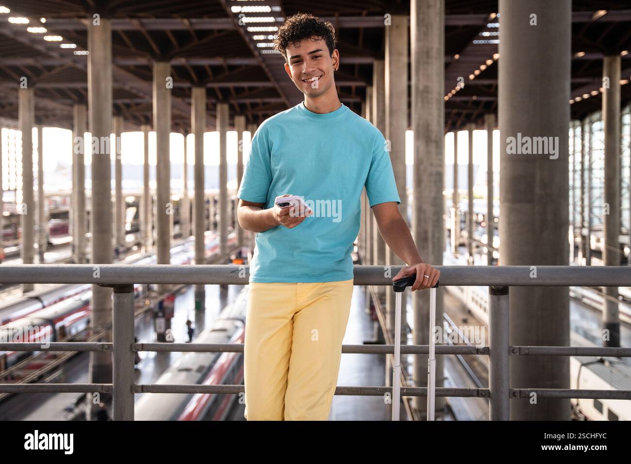 Un giovane viaggiatore con una camicia blu vibrante si appoggia casualmente contro una ringhiera in una stazione ferroviaria, controllando il suo telefono. Bagagli e treni sono visibili benea Foto Stock