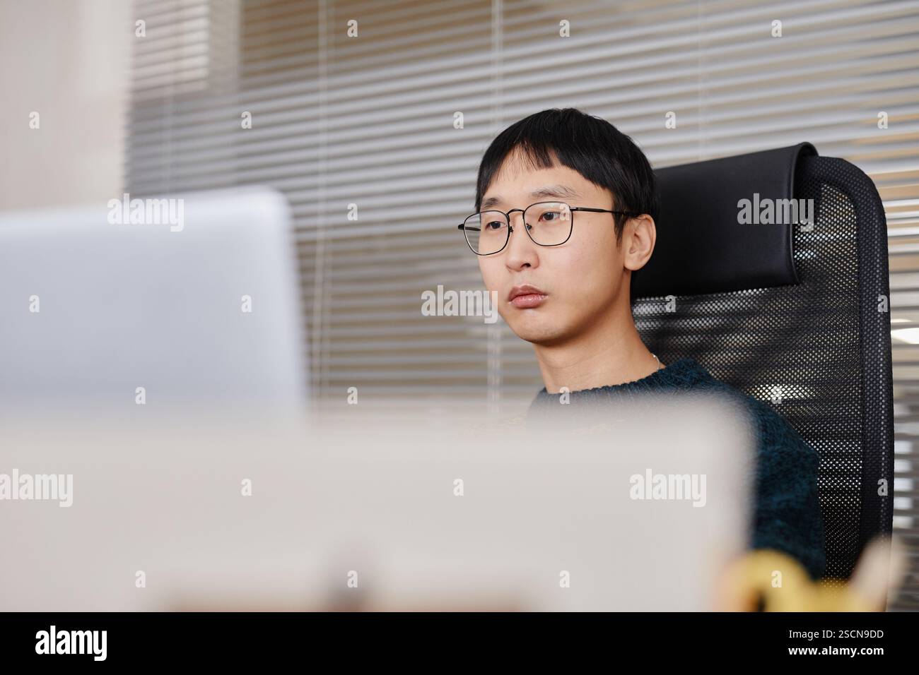 Ritratto di un giovane uomo asiatico che indossa gli occhiali, seduto alla scrivania dell'ufficio, che guarda lo schermo del computer in un moderno spazio ufficio con divisori in vetro Foto Stock