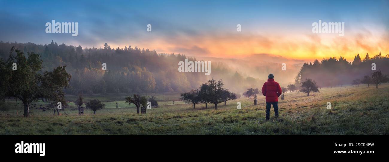 L'uomo in piedi su una collina nebbiosa all'alba guarda il bellissimo bagliore arancione in inverno. Un paesaggio panoramico girato nella campagna in Germania Foto Stock