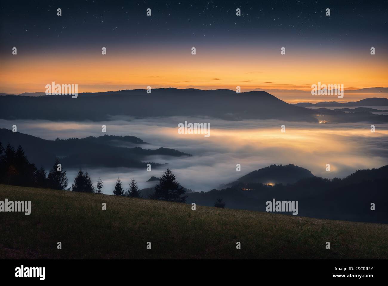 Paesaggio di montagna di notte sopra la nebbia nella valle, con bagliore dorato sull'orizzonte Foto Stock