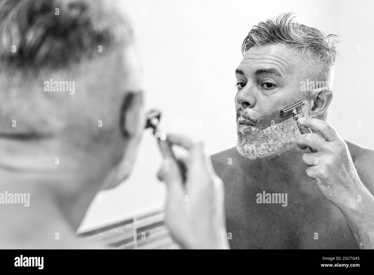 Uomo che si depone con un rasoio di sicurezza davanti allo specchio del bagno. La composizione in bianco e nero enfatizza la routine di cura del corpo, la cura di sé e i traditi Foto Stock