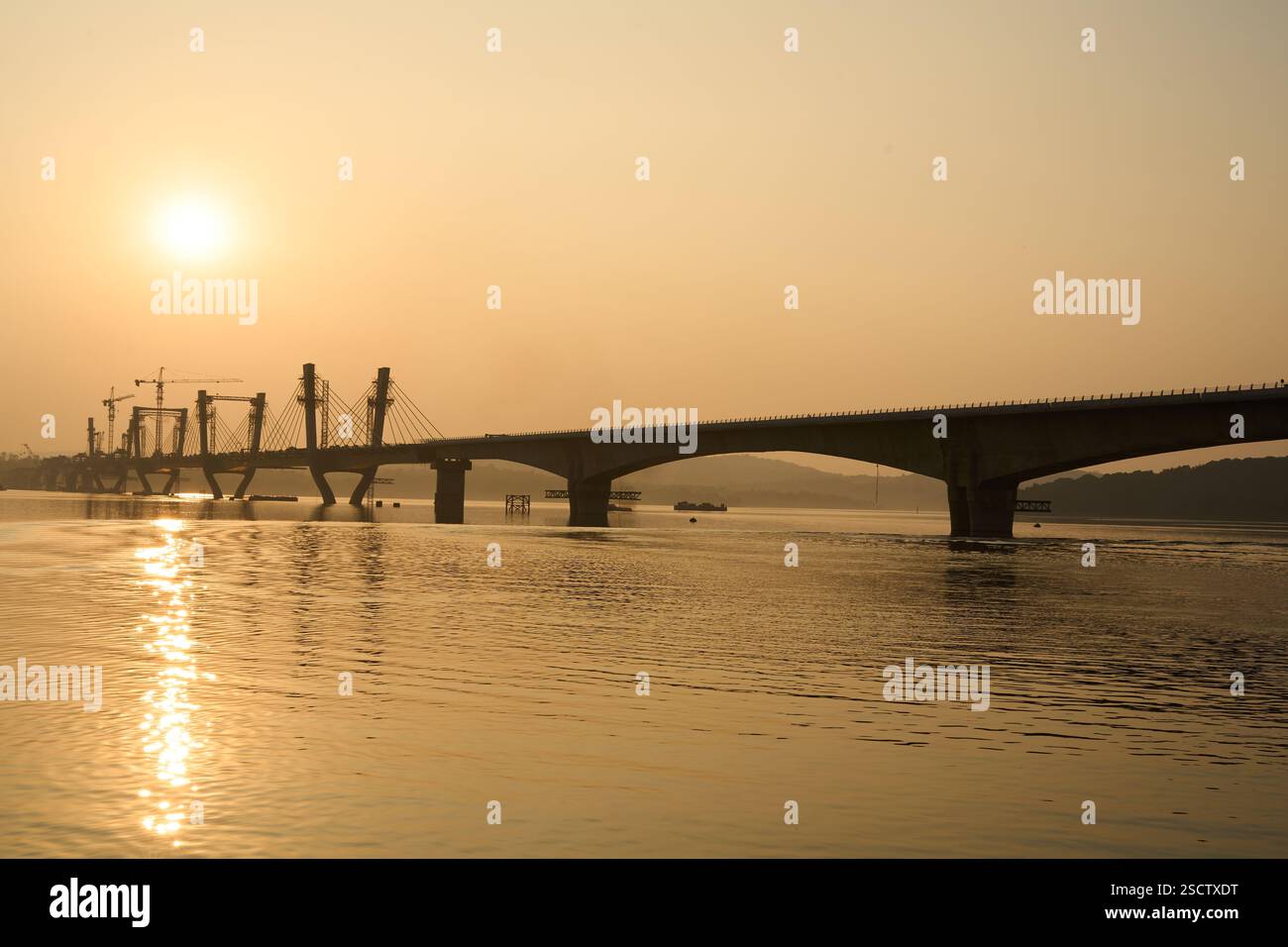 Una vista panoramica di un ponte strallato in costruzione al tramonto, con una luce dorata che si riflette sulle calme acque del fiume. Il tranquillo paesaggio controverso Foto Stock