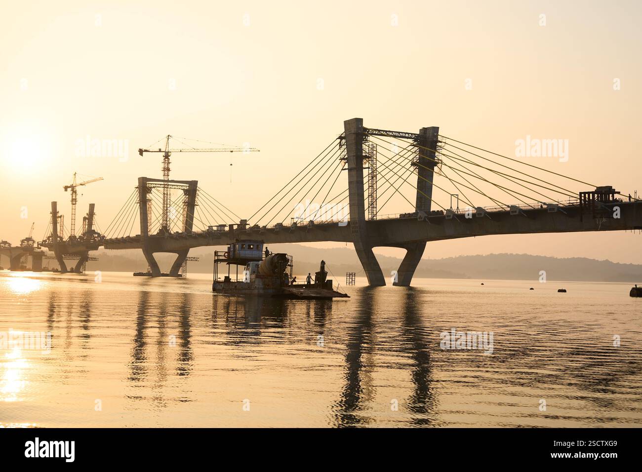 Una vista panoramica di un ponte strallato in costruzione al tramonto, con una luce dorata che si riflette sulle calme acque del fiume. Il tranquillo paesaggio controverso Foto Stock