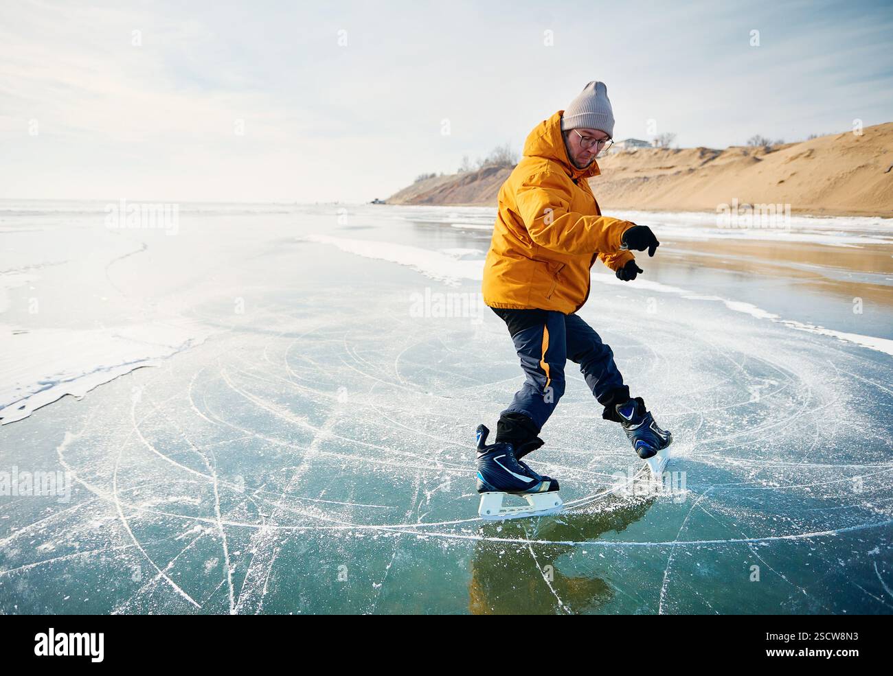 Atleta uomo in giacca gialla pattinaggio su ghiaccio sul lago ghiacciato con crepe e neve in inverno Foto Stock