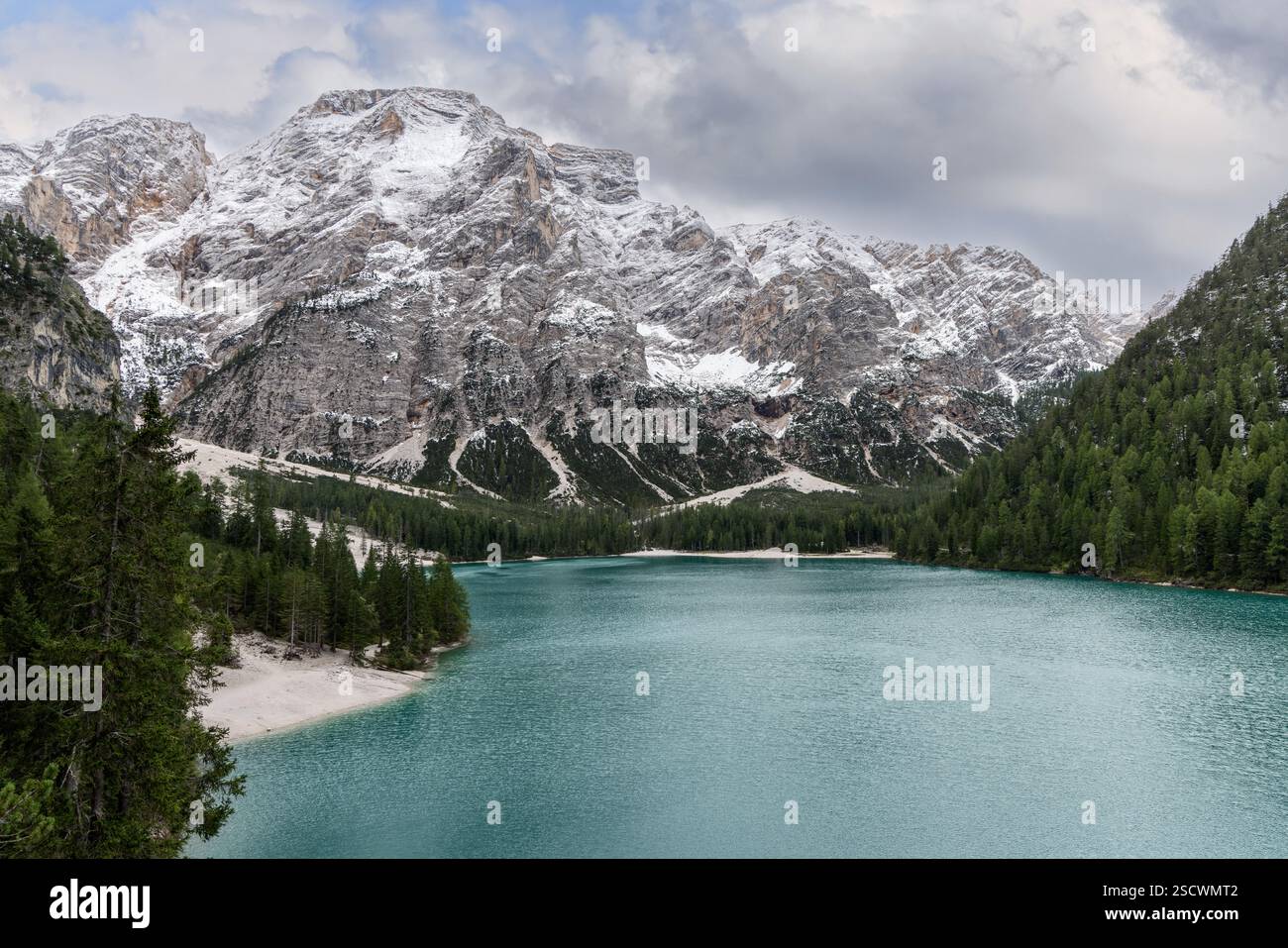 Lago Braies con acque turchesi circondate da fitte foreste sempreverdi e scogliere innevate sotto un cielo nuvoloso che forma una montagna mozzafiato Foto Stock