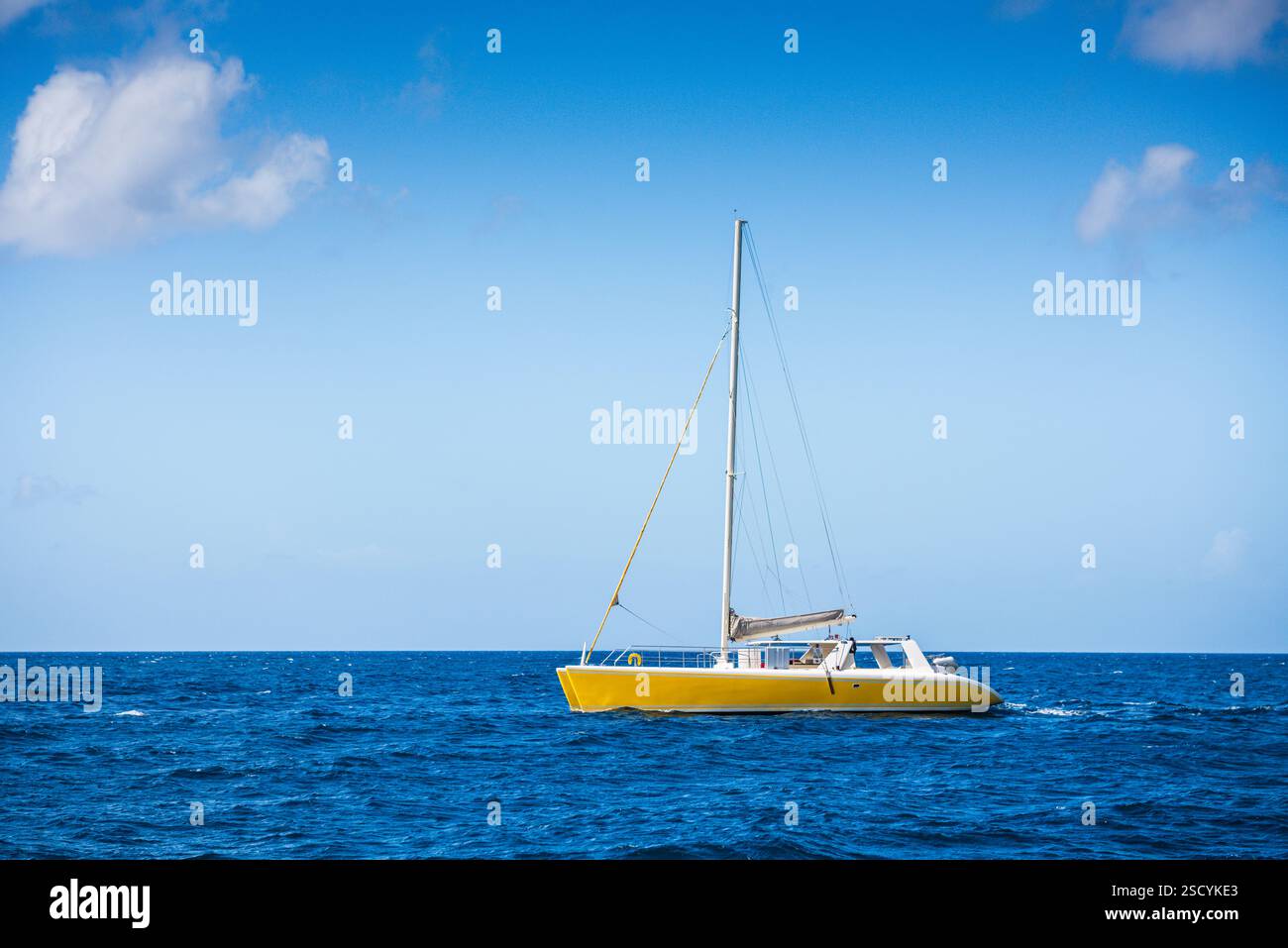 Catamarano giallo che naviga sul vivace blu del Mar dei Caraibi in una giornata di sole con piccole nuvole bianche che punteggiano il cielo blu. Foto Stock