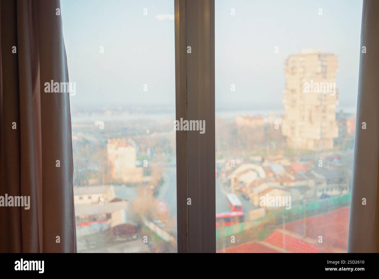 I dettagli di una finestra a doppia sospensione con vetro sporco, una tenda è visibile sul lato e i contorni degli edifici in lontananza attraverso la finestra. Foto Stock