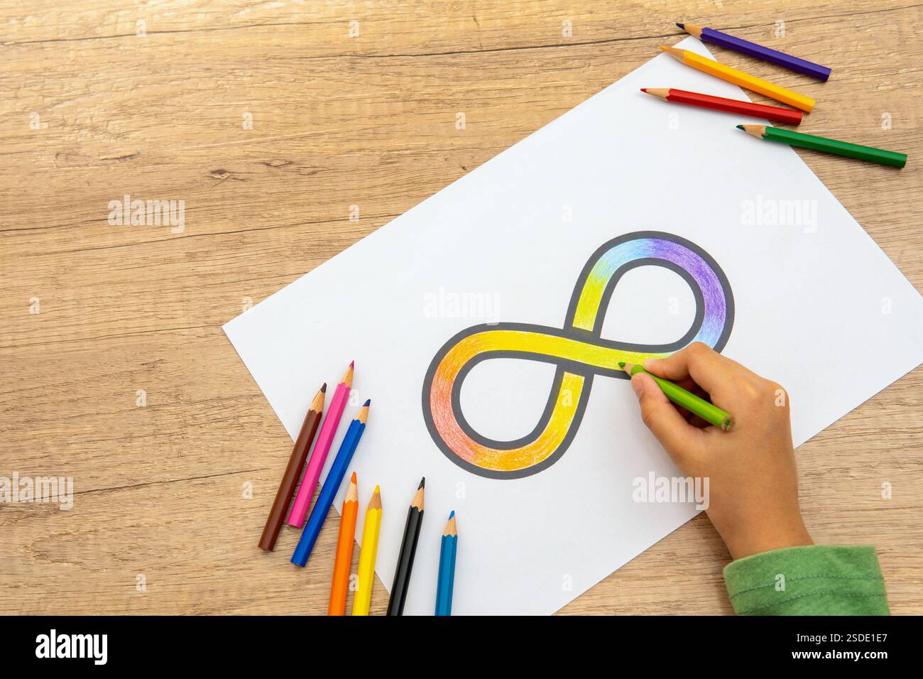 Una mano che incolla il simbolo dell'infinito arcobaleno su un foglio di carta bianco con matite colorate sparse intorno. Concetto di consapevolezza dell'autismo. Foto Stock