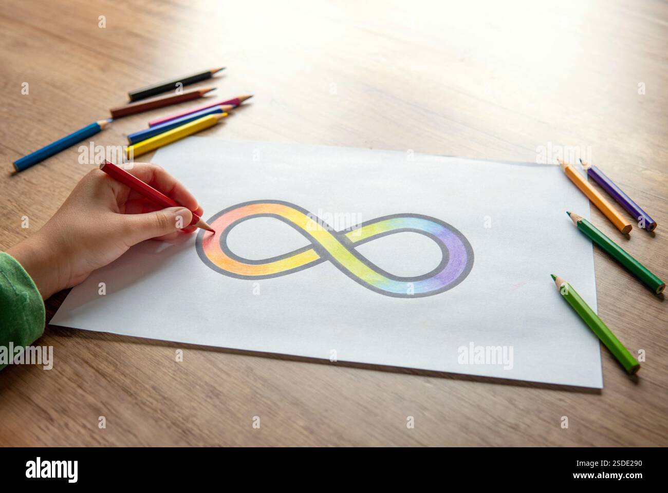 Una mano che incolla il simbolo dell'infinito arcobaleno su un foglio di carta bianco con matite colorate sparse intorno. Concetto di consapevolezza dell'autismo. Foto Stock
