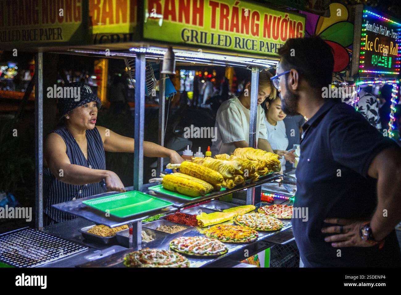 Un venditore di Street food interagisce con i clienti mentre serve cibo in un vivace mercato notturno di Hoi An, Vietnam, mercoledì 6 novembre 2024. Foto Stock