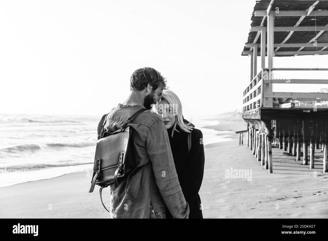 Romantica coppia hipster innamorata in piedi sulla spiaggia selvaggia vicino alla struttura in legno - fidanzato e fidanzata che indossano abiti alla moda e.. Foto Stock