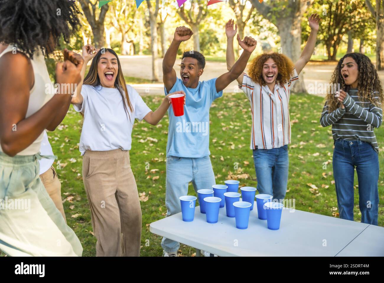Amici multietnici felici che alzano le braccia per celebrare un punteggio mentre giocano a beer pong in un parco Foto Stock