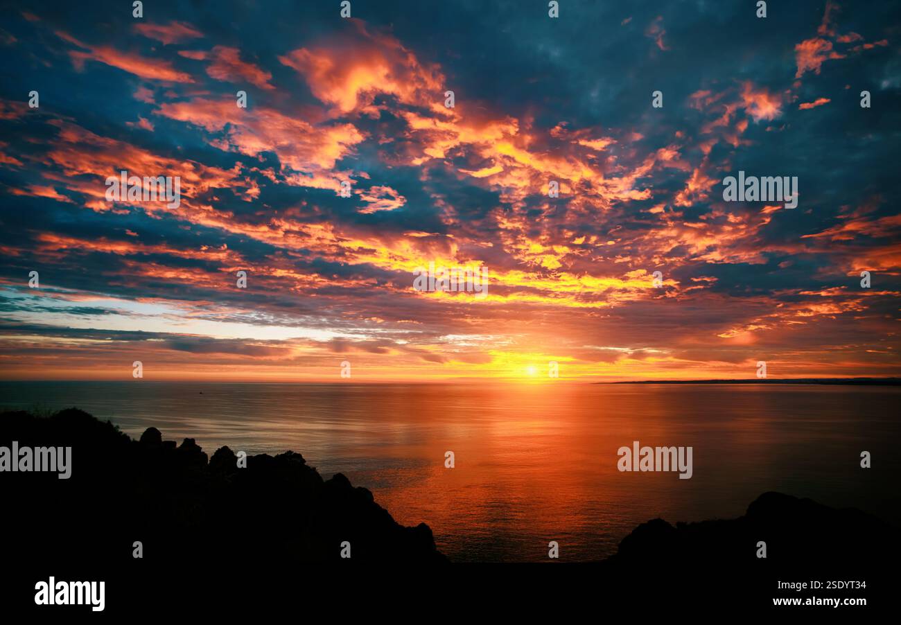 Spettacolare alba colorata sull'oceano con bellissime nuvole arancioni, sagome delle scogliere sulla costa e il sole riflesso sull'acqua Foto Stock