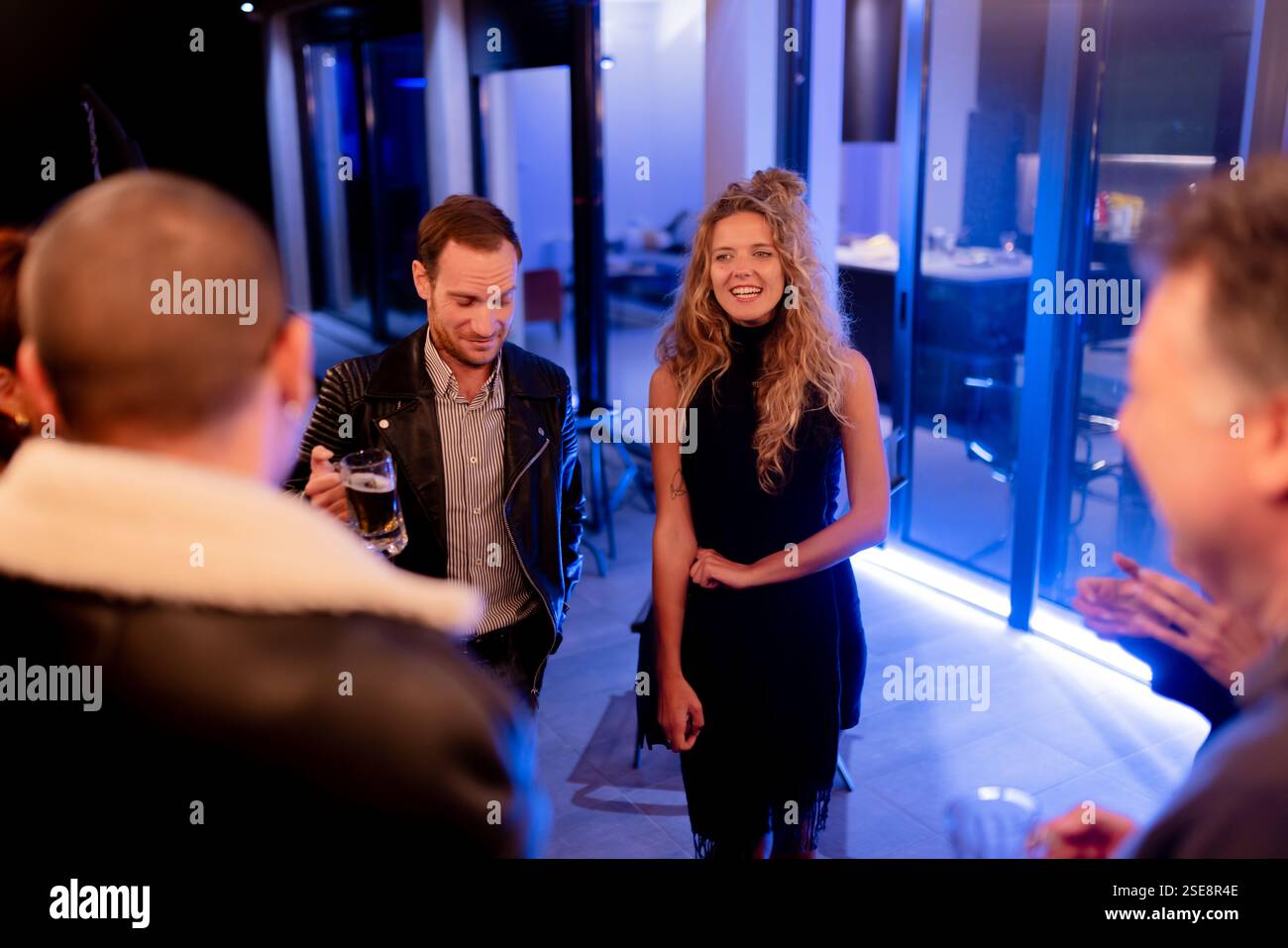 Un gruppo di persone che condividono un momento piacevole in un moderno raduno all'interno, vestito in festa e sorseggiando un drink sotto un'illuminazione elegante. Foto Stock