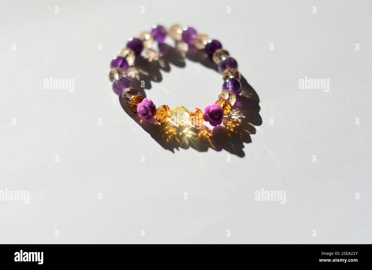 braccialetto con perline a forma di rosa viola, ambra e rosa, elegantemente visualizzato su sfondo bianco con riflessi di luce naturale Foto Stock