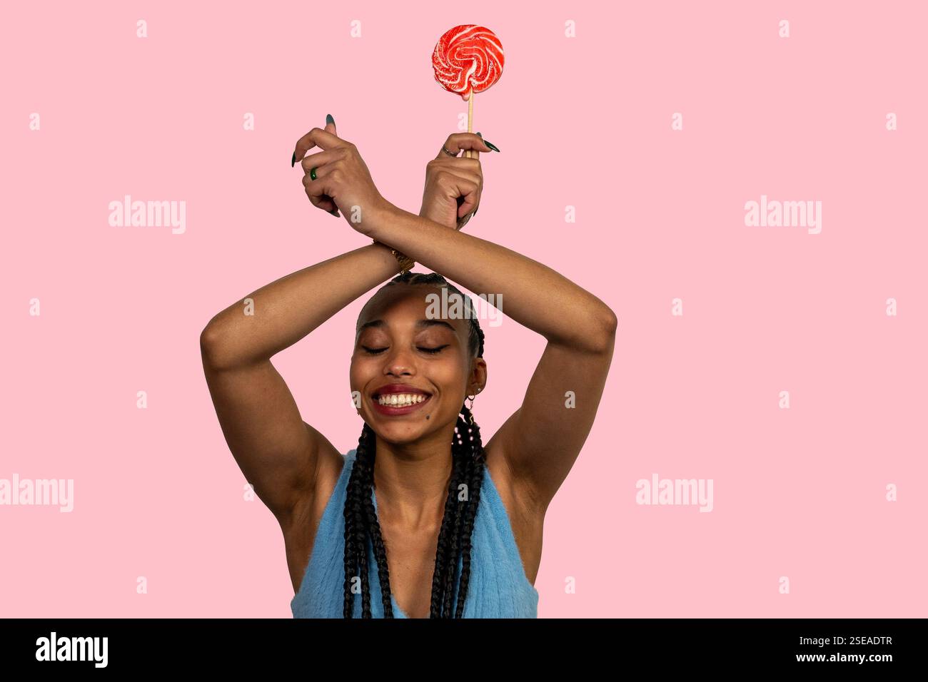 Giovane donna felice con trecce che reggono lecca rossa con braccia incrociate, sorridente e in piedi su sfondo rosa Foto Stock