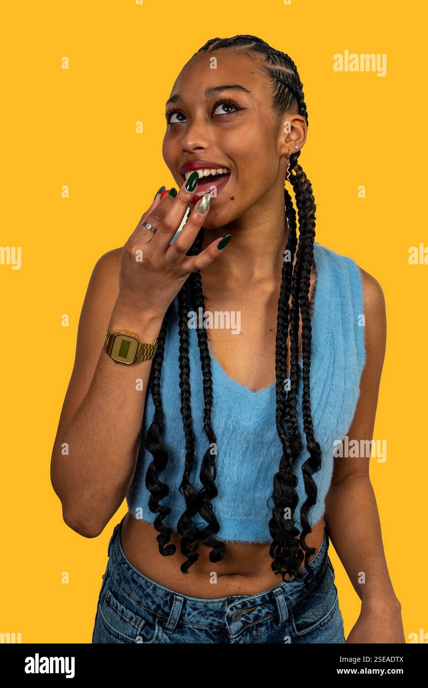 Giovane donna alla moda con lunghe trecce che si mordono le unghie con colorate nail art su un vivace sfondo giallo Foto Stock