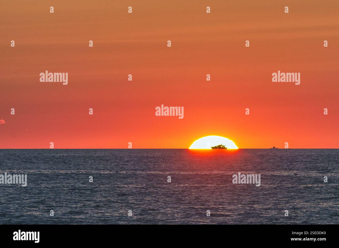 Un tramonto dorato sull'oceano con la silhouette di una nave all'orizzonte. Il sole si tuffa sotto la linea d'acqua, diffondendo tonalità calde attraverso il cielo e il mare. Foto Stock