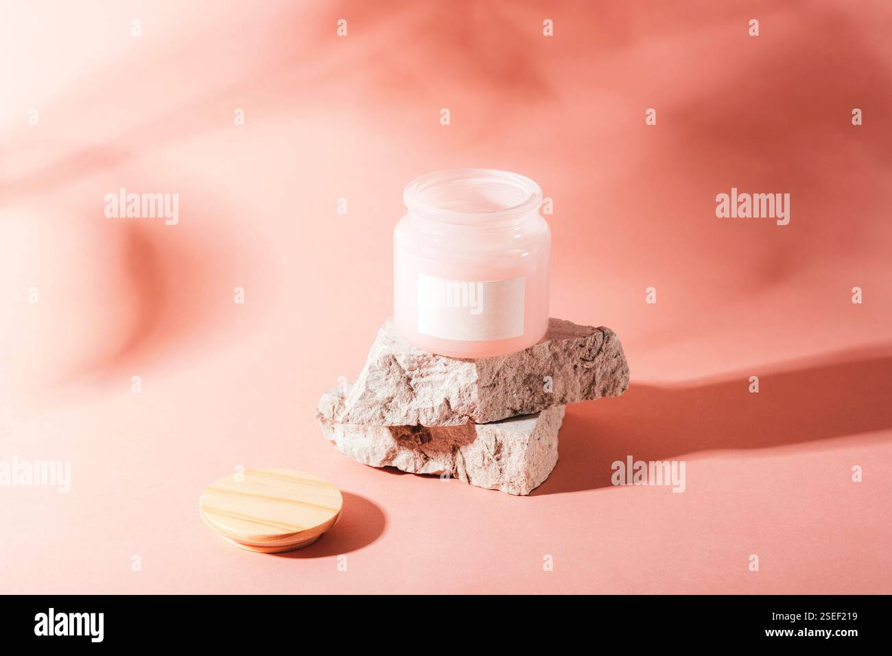 Apri il vaso per la cura della pelle con un coperchio in legno su pietre su sfondo rosa alla luce del sole. Mockup. Foto Stock