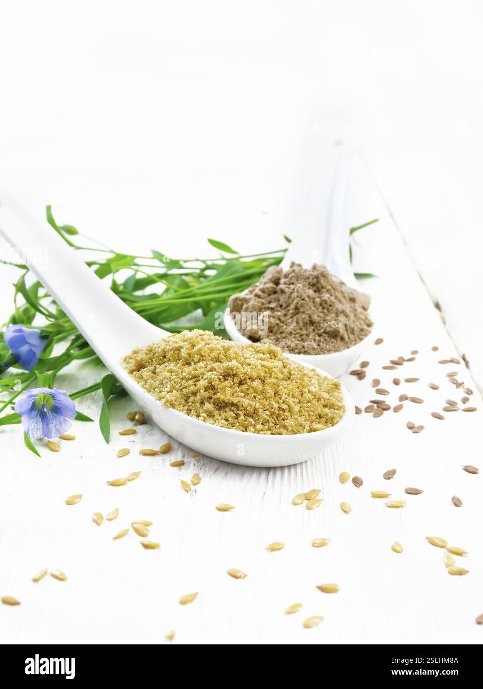 Crusca di lino e farina in due cucchiai, semi sul tavolo, foglie di lino e fiori sullo sfondo di tavole di legno chiaro Foto Stock Crusca di lino e farina in due cucchiai, semi sul tavolo, foglie di lino e fiori sullo sfondo di tavole di legno chiaro Foto Stock