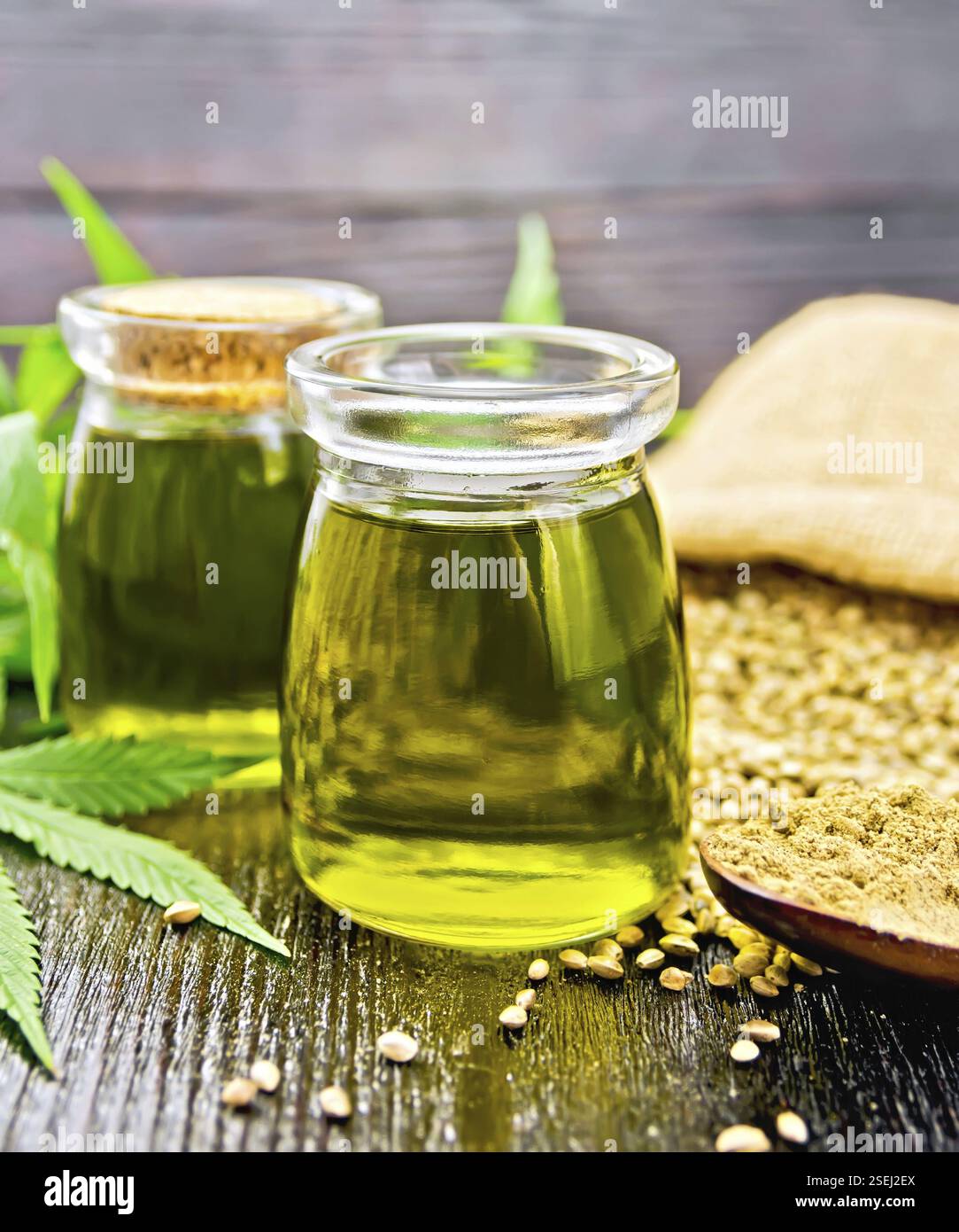 Olio di canapa in due vasetti di vetro, grano in un sacchetto e sul tavolo, farina in un cucchiaio, foglie di cannabis su uno sfondo di legno Foto Stock