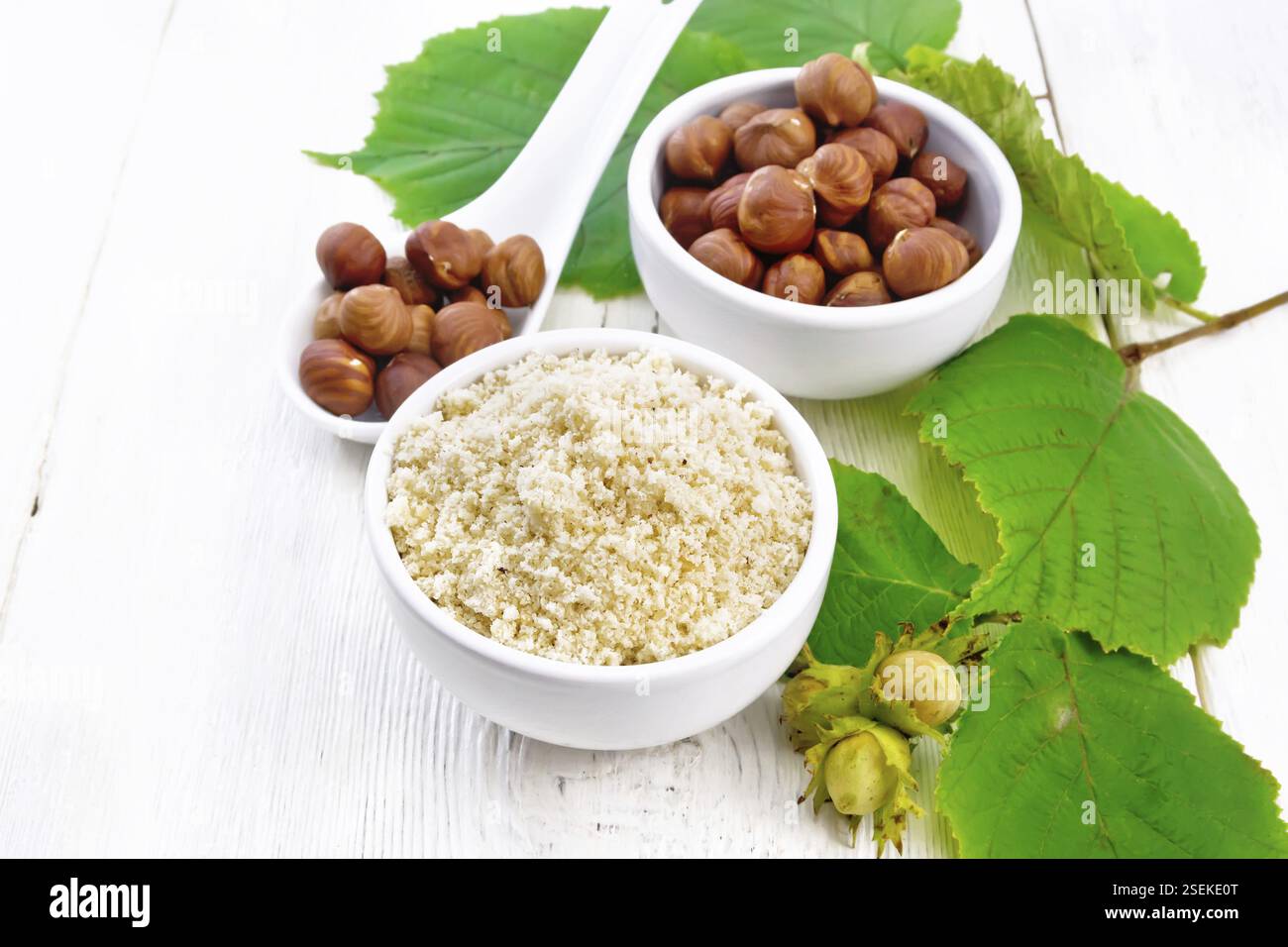 Farina e nocciole in due ciotole, un cucchiaio con nocciole pelate e un ramo di filbert con foglie sullo sfondo di legno chiaro Foto Stock