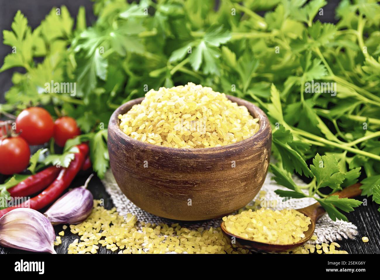 Bulgaro groats - grani di grano al vapore - in una ciotola di argilla e un cucchiaio su insaccatura, pomodori, peperoni caldi, aglio e prezzemolo su backgrou di legno nero Foto Stock