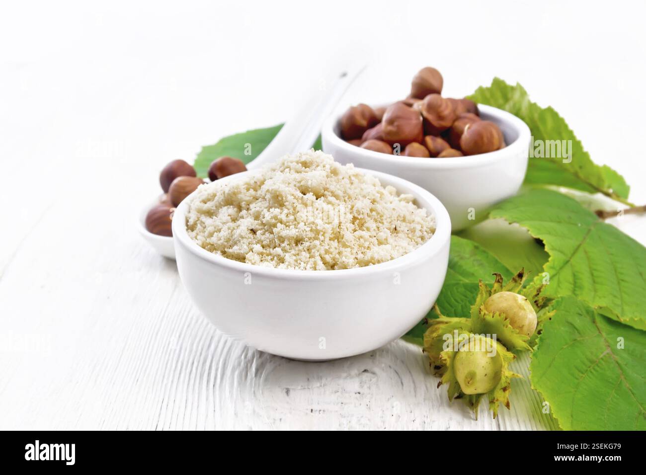 Farina e nocciole in due ciotole, un cucchiaio con nocciole pelate e un ramo di filbert con foglie verdi sullo sfondo di legno chiaro Foto Stock