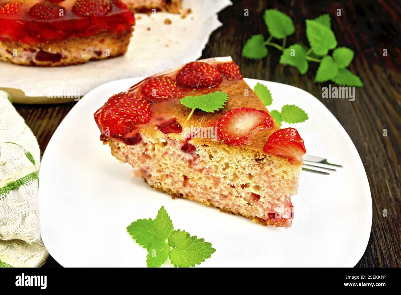 Pezzo di torta dolce con fragole, kissel, gelatina e menta, forcella in piastra bianca, tovagliolo contro lo sfondo delle tavole di legno Foto Stock