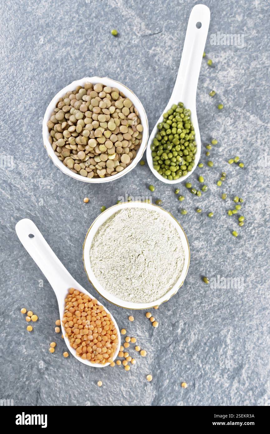 Farina di lenticchie in ciotola, lenticchie rosse e verdi in cucchiai, marrone in ciotola, gambi di fagioli con foglie sullo sfondo della tavola di pietra dall'alto Foto Stock